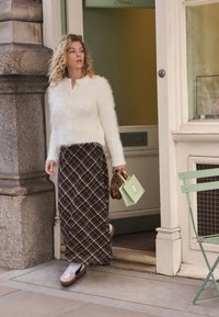 Cardigan blanc en tissu pelucheux, jupe longue à carreaux marron, baskets blanches avec des accents noirs, et petit sac à main marron. Sac de courses vert à la main.