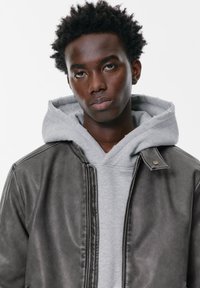 Giovane uomo di colore con capelli ricci corti, che indossa un felpa grigia sotto una giacca di pelle nera, guarda direttamente davanti a sé con un'espressione neutra.