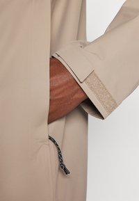 Bežs žakets ar teksturētu piedurkni un velcro aizdari, ar regulējamu auklu, kas ir melnbaltā pinumā.