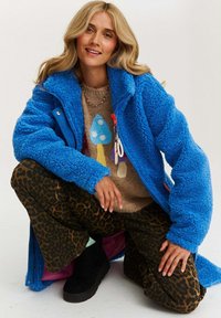 Blondínka sa zohýba, oblečená v jasnej modrej fleecovej bunde, svetri s hubovým vzorom, nohaviciach s leopardím vzorom a čiernych platformových čižmách.