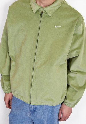 Osoba nosząca jasną zieloną sztruksową kurtkę na zamek z kołnierzem i małym białym logo Nike, zestawioną z jasnoniebieskimi jeansami.