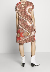 Žena v červených vzorovaných šatech po kolena s vykrojenými detaily na horní části zad a bílých nákladních botách na podpatku, stojící na šedé podlaze.