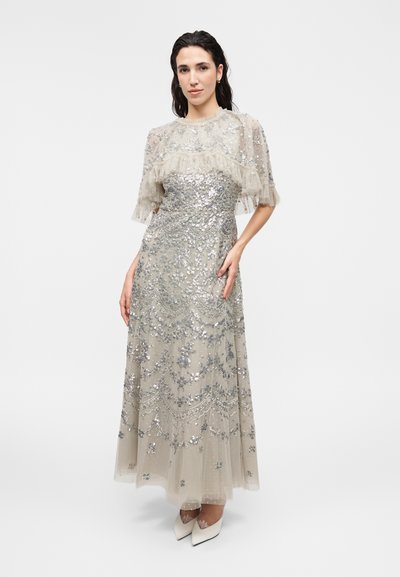 Γυναίκα που φοράει ένα ασημένιο μακρύ φόρεμα με παγέτες και δαντέλα, διάφανα μανίκια με βολάν και λευκά μυτερά τακούνια, στέκεται μπροστά σε λευκό φόντο.
