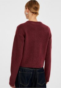 Burgundy kortärmad tröja i mjukt, texturerat tyg med rund halsringning och långa ärmar, i kombination med mörk denim.