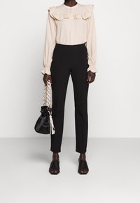 Blouse beige à volants avec manches longues, pantalon noir taille haute et ajusté, sandales noires à brides, et sac à cordon noir avec des accents crème.
