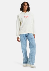 Sudadera beige clara con un bolsillo frontal, que presenta un pequeño gráfico en rojo y amarillo. Combinada con unos vaqueros de pierna ancha azul claros y zapatillas blancas.