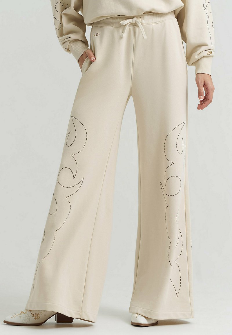 Beige wijde broek met trekkoord in de taille, decoratieve geborduurde vlammenpatronen op de benen, gedragen met een bijpassende beige top en laarzen met hoge hak en bloemenborduursel.