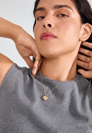 Young person with smooth skin wearing a gray sleeveless top and a gold heart-shaped pendant necklace, hands touching neck and head.