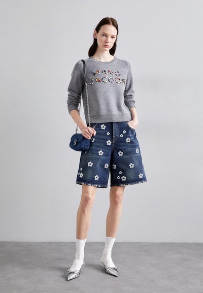 Suéter gris con logotipo decorativo, combinado con pantalones cortos de denim azul con bordado floral blanco. Zapatos plateados de punta y calcetines blancos.