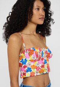 Top corto floreale con spalline sottili, caratterizzato da vivaci fiori arancioni, rosa e blu su uno sfondo bianco. Design del busto strutturato.