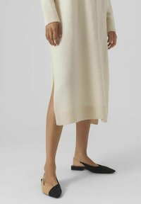 Robe crème à manches longues avec une fente sur le côté, fabriquée en matériau doux. Chaussures slingback noires et beige avec des bouts pointus et de petits talons carrés.