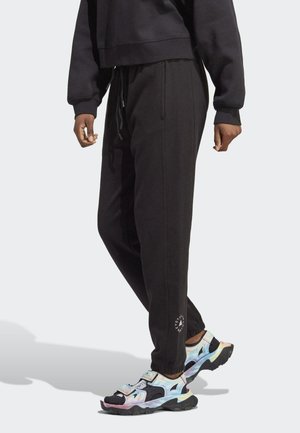Pantalon de jogging noir avec taille à cordon de serrage, chevilles resserrées, poches latérales et un logo circulaire près de l'ourlet. Associé à des chaussures de sport multicolores.