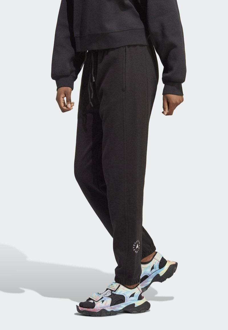 Schwarze Jogginghose mit einem Kordelzugbund, mit Bündchen an den Knöcheln, seitlichen Taschen und einem runden Logo nahe dem Saum. Kombiniert mit mehrfarbigen Sportschuhen.