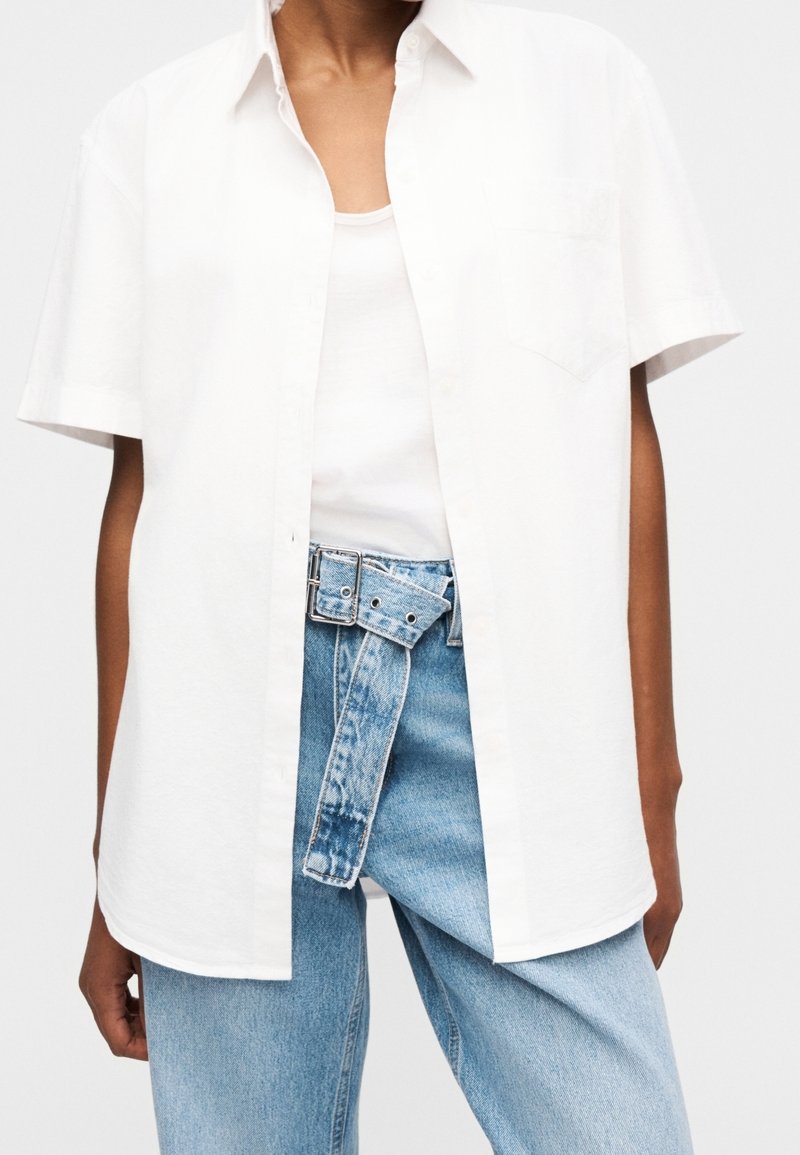 Personne portant une chemise blanche à manches courtes boutonnée sur un débardeur blanc et un jean taille haute bleu clair avec une large ceinture en jean.