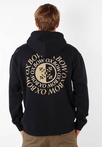 Hoodie noir en mélange de coton, présentant un grand logo doré et un texte au dos. Coupe décontractée avec poche kangourou et capuche à cordon.