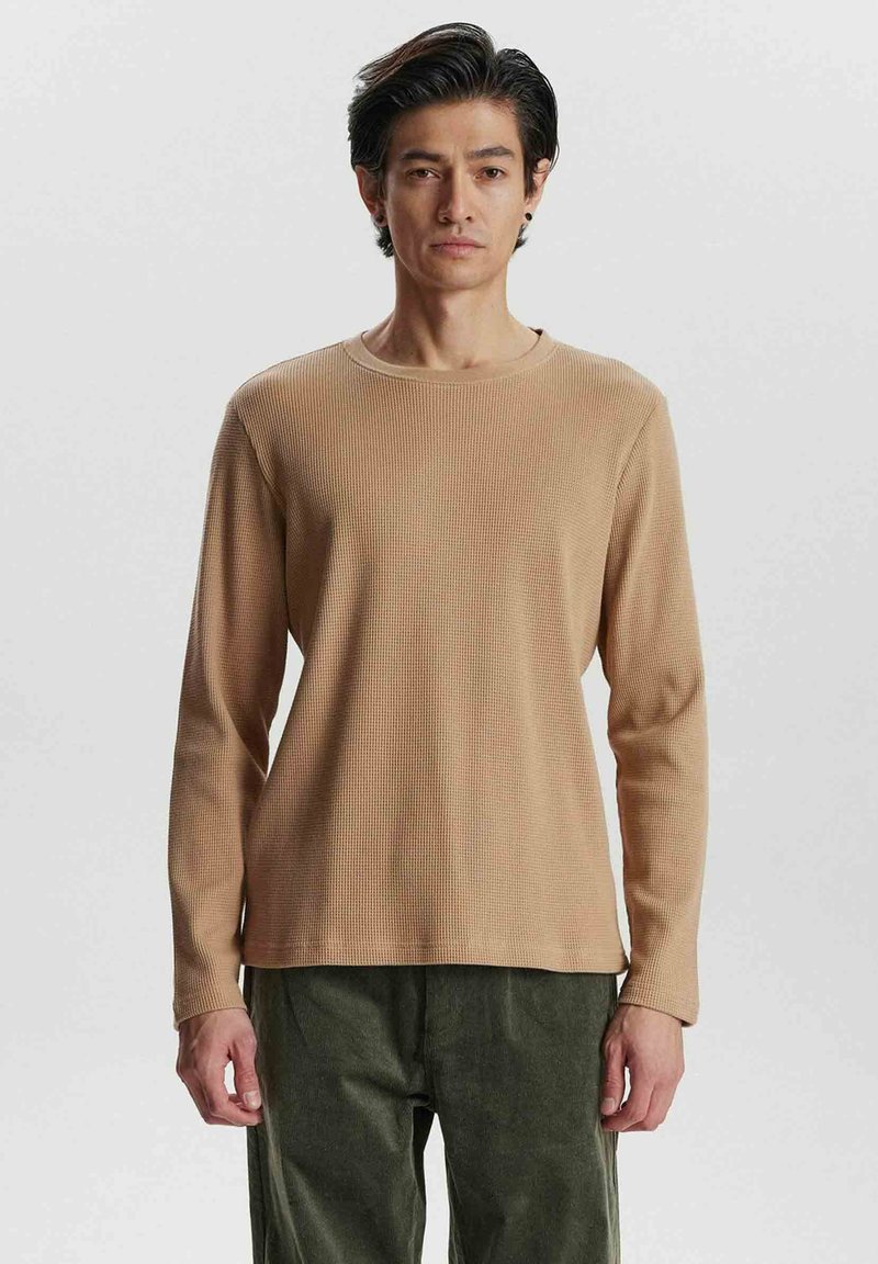 Beige lange mouwen ribbelshirt met een ronde halslijn. Zachte textuur, rechte snit, gedragen met donkergroene corduroy broek.