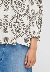 Hvid bluse med brun blomsterbroderi, lange ærmer med samlede manchetter og løs pasform; båret over en blå denimnederdel.