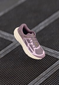 Svijetlopurpureta atletska cipela s mrežastim gornjim dijelom, kontrastnim teksturiranim preklopima i utezima s mekanom podlogom. Sadrži vezice i minimalne detalje brendiranja.