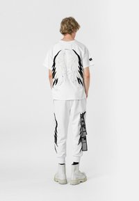 Camiseta blanca con bordado de alas en negro y plata en la parte posterior, combinada con pantalones jogger blancos que presentan detalles en negro y texto. Calzado casual.