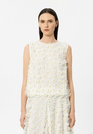 Junge Frau trägt ein ärmelloses weißes Top und Hosen, die mit weißen und gelben Gänseblümchen-Applikationen verziert sind, steht vor einem einfarbigen Hintergrund.