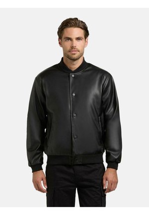 Man met kort haar, gekleed in een zwarte leren bomberjack met knopen en een zwarte broek, staand tegen een effen lichtgrijze achtergrond.