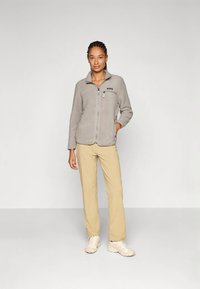 Helehall fleece-tagi, millel on esil lukk, küljetaskud ja rinnal logo. Ühitatud beeþide pükste ja heledate tossudega.