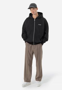 Haut à capuche zippé noir avec poches avant, accents gris et logo "PEGADOR". Associé à un pantalon plissé beige et des baskets blanches.