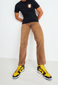 Svart T-shirt med kort ärm som har en färgglad logotyp, i kombination med beige raka byxor och svarta/gula high-top sneakers.