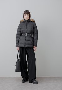 Vrouw staat en draagt een zwarte donzen jas met ceintuur en bontkraag, wijde zwarte broek, zwarte schoenen en houdt een zwarte handtas vast.