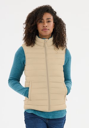 Kvinde med krøllet hår iført en beige quiltet vest over en blå langærmet skjorte, hænder i vestlommer, stående mod en hvid baggrund.