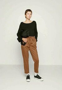 Top preto de mangas longas, calças castanhas risca de giz, meias brancas até ao tornozelo e sapatilhas pretas. O modelo segura uma jaqueta preta acolchoada. Fundo simples.