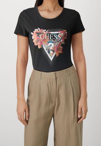 Žena v černém tričku Guess s květinovým a trojúhelníkovým logem, spárovaném s béžovými kalhotami s vysokým pasem, stojící na neutrálním pozadí.