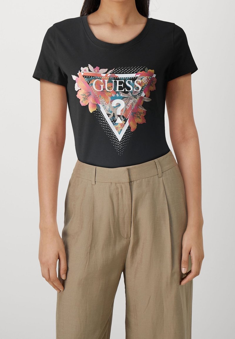Žena v černém tričku Guess s květinovým a trojúhelníkovým logem, spárovaném s béžovými kalhotami s vysokým pasem, stojící na neutrálním pozadí.