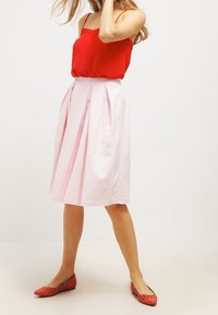 Gonna rosa plissettata al ginocchio in tessuto leggero, abbinata a una maglia rossa brillante. Indossata con ballerine rosse traforate.