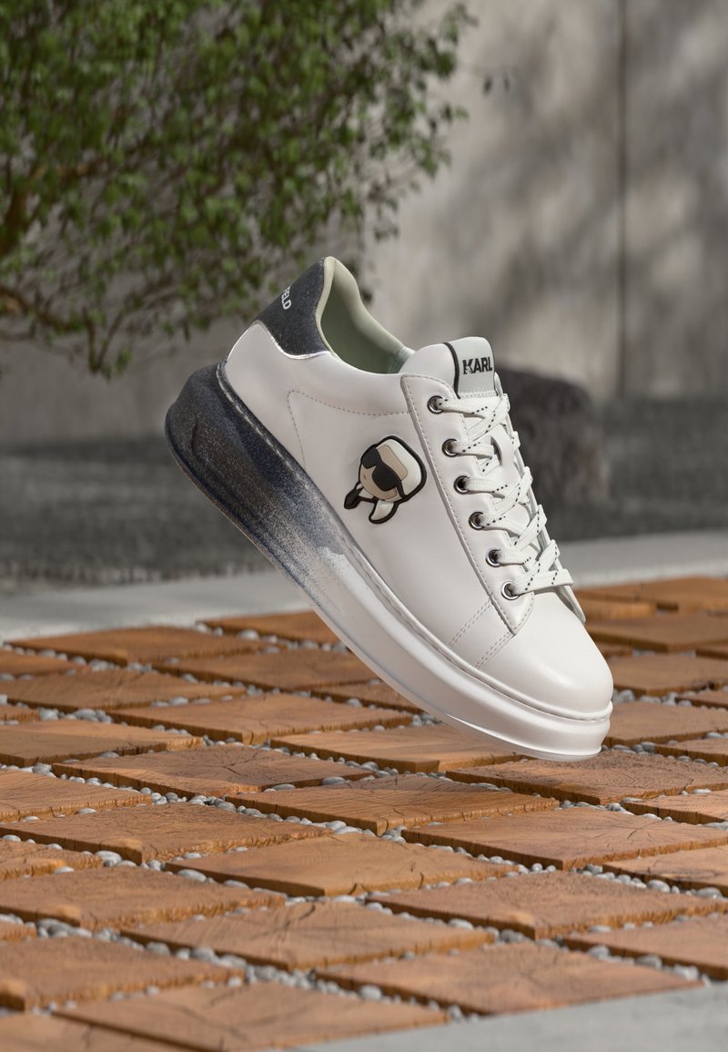 Λευκό δερμάτινο αθλητικό παπούτσι με γκρι και μαύρη σόλα, που φέρει ένα καρτούν λογότυπο στο πλάι και κορδόνια, παρουσιάζοντας μια λεία υφή και μινιμαλιστικό σχεδιασμό.
