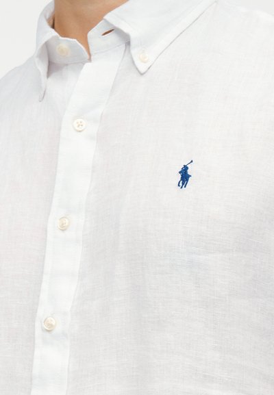 Λευκό πουκάμισο με κουμπιά και γιακά, με μπλε κεντημένο λογότυπο παίκτη πόλο στο αριστερό στήθος.