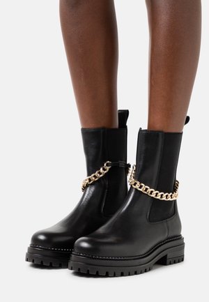 Botas de cuero negras hasta la mitad de la pantorrilla con suelas gruesas y detalle de cadena dorada, llevadas sobre piernas desnudas contra un fondo blanco.