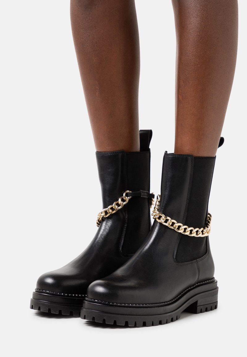 Czarny skórzany botki do połowy łydki z grubą podeszwą i złotym łańcuchem noszone na nagich nogach na białym tle.