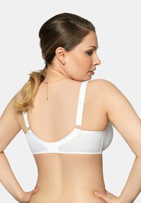 Donna con pelle chiara che indossa un reggiseno bianco, di spalle, con i capelli raccolti e le mani sui fianchi su uno sfondo semplice.