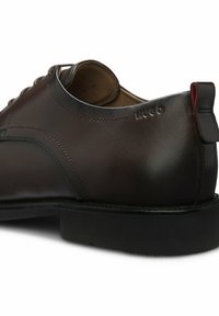 Dunkelbrauner Lederschuh mit Prägung "HUGO" nahe der Ferse und schwarzer Sohle mit hinterem Zuglasche.