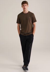 Hnědé tričko s krátkým rukávem a kulatým výstřihem, doplněné černými kalhotami s volným střihem. Obuv tvoří černé kožené mokasíny.