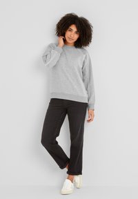 Woman with curly hair wearing a grey sweatshirt, black rolled-up jeans, and white sneakers, standing against a light grey wall.