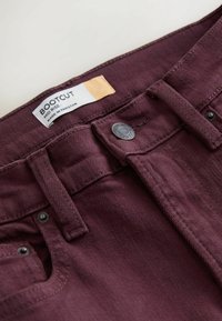 Nærbillede af bordeaux bootcut-jeans, der viser linning, knap, bæltestropper, nitter og et mærke med teksten "Bootcut Mid Rise Made in Pakistan."