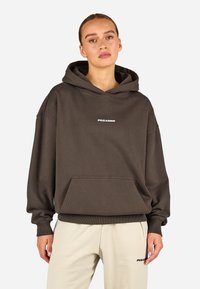 Brązowa, oversize bluza z kapturem z miękkiego materiału. Posiada przednią kieszeń kangurzą oraz białe logo na piersi. Rękawy i dół wykończone ściągaczem.