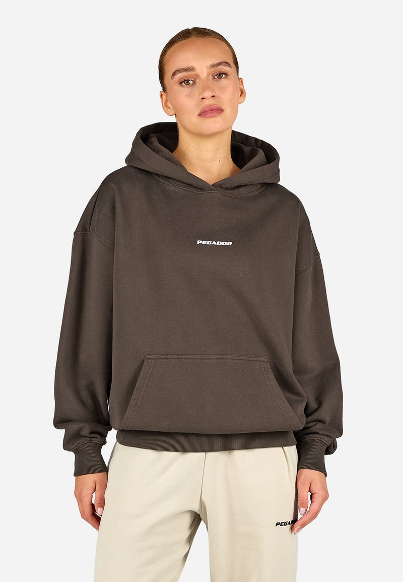 Brązowa, oversize bluza z kapturem z miękkiego materiału. Posiada przednią kieszeń kangurzą oraz białe logo na piersi. Rękawy i dół wykończone ściągaczem.