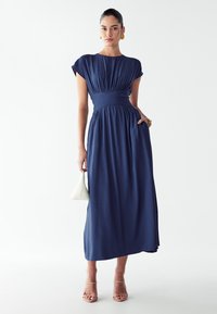 Mornarsko modra maxi obleka s kratkimi rokavi, nabranim trupom in prilegajočim pasom, ki ima tekočo krilo in je obuta v nagubane nude sandale.