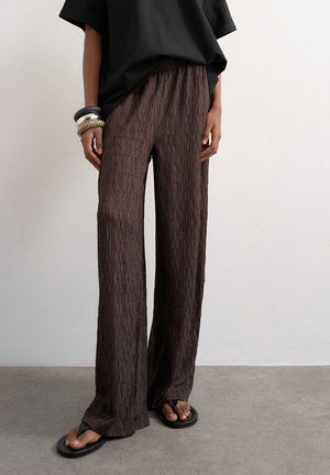 Persoon die textured bruine wijd uitlopende broek draagt, zwart oversized top, zwarte slippers en meerdere armbanden aan de linkerpols.