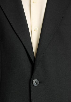 Melnā pielāgotā uzvalka žakete ar teksturētu apdari, kas satur vienu pogu un kontrastējošu gaišas krāsas krekls ar redzamām pogām.