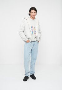 Sweatshirt à capuche gris avec doublure en peluche, motif graphique ; jean en denim bleu clair ; chaussures noires ; debout sur un fond uni.