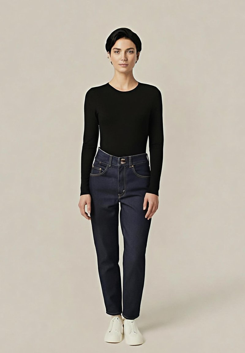 Haut noir à manches longues associé à un jean taille haute bleu foncé. Le jean présente des poches avant et une fermeture à bouton. Le modèle porte des chaussures blanches.